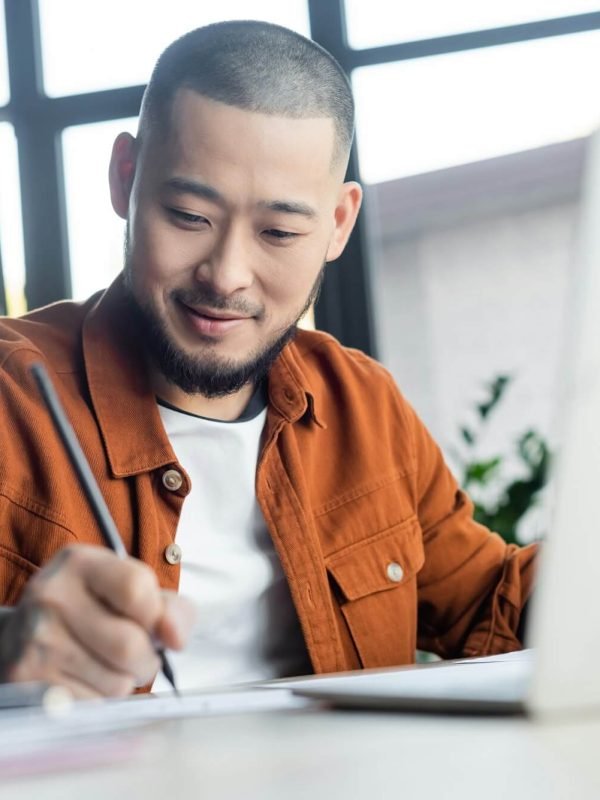 smiling-asian-designer-drawing-near-blurred-laptop-in-architectural-bureau.jpg